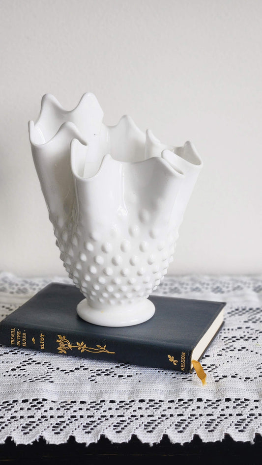 Vintage Fenton hobnail milk glass handkerchief vase with ruffled top displayed on vintage book on lace cloth